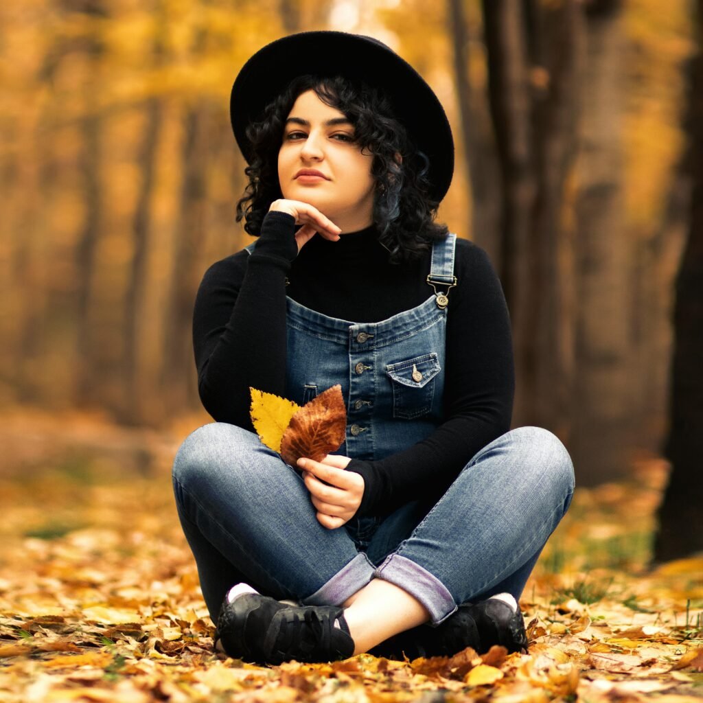 A young woman in denim overalls and a hat poses amidst autumn leaves in Tabriz, Iran.